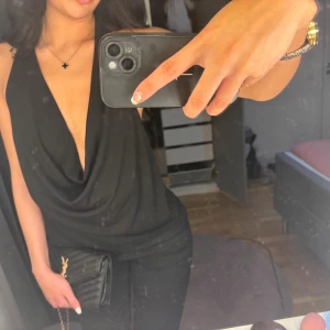 Svart omlott halterneck topp - Säljer en svart omlott halterneck topp med djup v-ringning och öppen rygg. Toppen har en snygg draperad framsida och är ärmlös, perfekt för att skapa en stilren och trendig look. Materialet känns mjukt och följsamt mot huden.