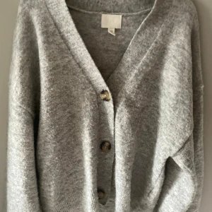 Grå kofta från H&M med knappar - Mysig grå kofta från H&M med v-ringning och tre stora knappar framtill. Koftan är stickad och har en loose passform med långa ärmar. Perfekt för lager-på-lager och chill dagar. Enkel och stilren design som funkar till det mesta.