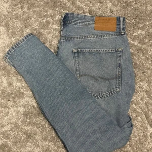 Jack an Jones ljusblå jeans -  jeans i ljusblå denim med rak passform och hög midja. Byxorna har fem fickor, synlig söm på bakfickan och en brun läderpatch med logga bak i midjan. Perfekta för en avslappnad och tidlös look.