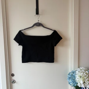 Svart offshoulder croppad topp M - Säljer en svart croppad topp från Monki med offshoulder-design och korta ärmar. Enkel och stilren modell som är perfekt att matcha med höga jeans eller kjol. Toppens material känns mjukt och stretchigt för en bekväm passform.