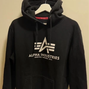 Svart hoodie från Alpha Industries - Svart hoodie från Alpha Industries med vit logga och text på bröstet. Tröjan har huva med snörning och en stor magficka framtill. Perfekt för dig som gillar streetwear och vill ha en stilren look.