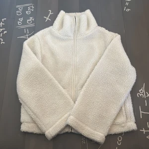 Vit teddyjacka med dragkedja - Mysig vit teddyjacka med hög krage och hel dragkedja framtill. Jackan har två stora fickor och är tillverkad i fluffigt teddy-material som ger en riktigt skön och avslappnad vibe. Perfekt för kyliga dagar när du vill vara både varm och stilren. 1 gång använd