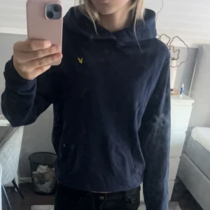Marinblå hoodie från Lyle & Scott - Snygg marinblå hoodie från Lyle & Scott med klassisk gul logga på bröstet. Tröjan har huva, känguruficka och är tillverkad i mjuk bomull. Perfekt för dig som gillar en enkel och stilren look. Nypris: 749kr
