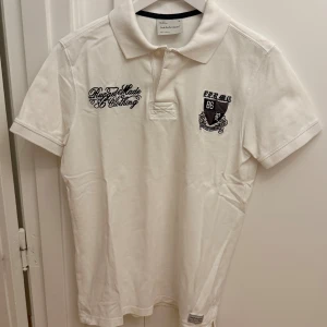 Vit pikétröja Peak Performance M - Vit pikétröja från Peak Performance i 100% bomull. Klassisk krage, korta ärmar och broderade detaljer på bröstet med logga och text. Perfekt för en clean och sportig stil. Snygg passform och stilren look. Storlek M.