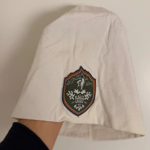 Vit mössa från Muddus med Fjällgården-märke - Vit mössa från Muddus med ett stort broderat Fjällgården-märke framtill. Mössan är tillverkad i mjuk bomull med lite stretch för skön passform. Perfekt för dig som gillar outdoor och vill ha en clean stil med en touch av fjällkänsla.