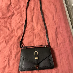 Svart axelväska med gulddetaljer - Stilren svart axelväska i syntetmaterial med justerbar rem och snygga guldfärgade metalldetaljer. Väskan har ett kuvertformat lock och dekorativt spänne framtill. Mycket bra utrymme