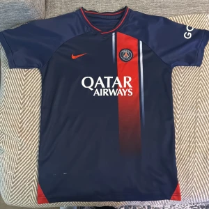 PSG matchtröja Nike blå/röd - Paris Saint-Germain fotbollströja från Nike i marinblått med röd vertikal rand och vita detaljer. PSG-logga på bröstet, Qatar Airways tryck och orange Nike swoosh. Kortärmad modell i lätt, glansigt material som andas. Perfekt för dig som älskar fotboll och PSG.