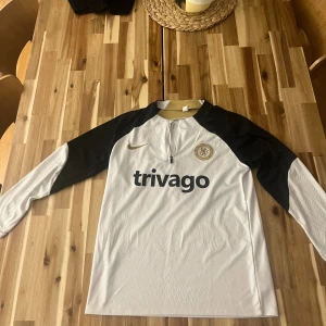 Chelsea långärmad tränings-topp Nike - Chelsea FC långärmad tränings-tröja från Nike i vitt med svarta ärmar och guldiga detaljer vid kragen. Tröjan har halv dragkedja framtill, broderad Nike-logga och Chelsea-märke på bröstet samt 'trivago' tryckt på mitten. Perfekt för träning och hösten