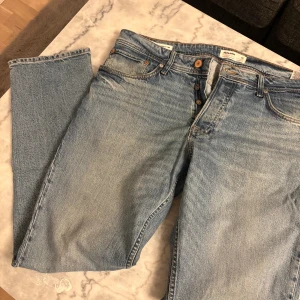 Ljusblå jeans från Jack & Jones - Säljer ett par klassiska ljusblå jeans från Jack & Jones med raka ben och femficksdesign. Jeansen har knäppning med knappar och snygga kontrastsömmar. Perfekta för en avslappnad och stilren look.