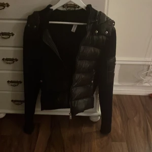 Svart dunjacka från Moncler - Svart Moncler dunjacka med quiltad framsida och huva. Jackan har långa ärmar, dragkedja och Moncler-logga på ärmen. Materialmix med dun och stickade partier ger en modern look. Perfekt för kyliga dagar.