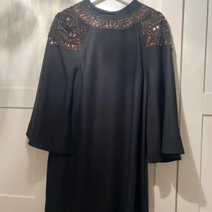 Svart klänning med paljettdekor Spotlight - Svart kort klänning från Spotlight by Warehouse med vida trekvartsärmar och snygga paljett- och pärldetaljer på axlar och runt halsringningen. Klänningen har en rak passform och är perfekt för dig som vill sticka ut med coola detaljer.