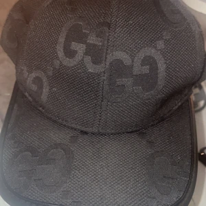 Svart keps från Gucci med GG-mönster - Snygg svart keps från Gucci med diskret GG-monogram över hela kepsen. Tillverkad i tåligt tyg med klassisk böjd skärm och detaljer i svart. 