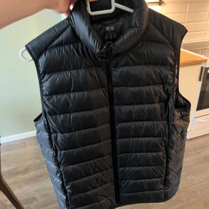Svart dunväst från Uniqlo M - Svart, ultralätt dunväst från Uniqlo i storlek M. Västen har quiltad design, hög krage och dragkedja framtill. Tillverkad i glansig polyester med dunfyllning. Perfekt för lager-på-lager och enkel att packa ner i medföljande påse.