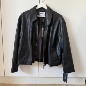 Svart skinnjacka från NA-KD, stl 36 - Snygg svart skinnjacka från NA-KD i storlek 36. Jackan har bred krage, raka fickor och dragkedja framtill. Tillverkad i syntetiskt skinn (PU) med en clean och stilren look. Perfekt för dig som vill ha en klassisk men trendig jacka.