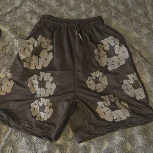 Svarta shorts från Denim Tears med tryck - Säljer ett par svarta shorts från Denim Tears med gråa, blommiga tryck över hela plagget. De har elastisk midja med snörning och är tillverkade i mjuk bomull. Perfekt för dig som gillar streetwear och unika prints.