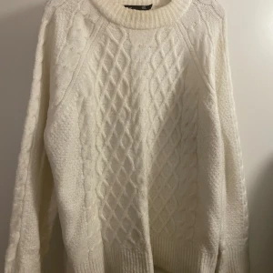 Vit stickad tröja från 157 - Knappt använd mysig vit stickad tröja från 157 med klassiskt flätmönster framtill och ribbade muddar. Tröjan har rund halsringning och långa ärmar, perfekt för kyliga dagar. Snygg och enkel att matcha med jeans eller kjol.