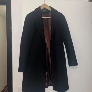 Svart kappa från Kappahl - Stilren svart kappa från Kappahl med klassisk krage och knäppning framtill. Fodret är glansigt och har en diskret randig effekt i vinröd ton. Långärmad och rak passform, perfekt för kyligare dagar.