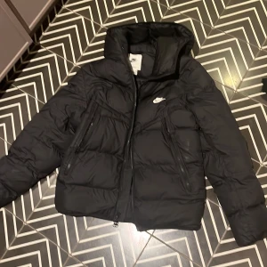 Svart vinterjacka från Nike - Svart pufferjacka från Nike Storm-FIT med huva och två snedställda dragkedjefickor framtill. Jackan har en klassisk pufferlook, diskret Nike-logga på bröstet och är tillverkad i syntetmaterial. Perfekt för kalla dagar när du vill hålla stilen.