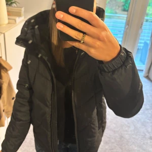 Svart pufferjacka från Barbour - Svart pufferjacka från Barbour International med huva och snygg quiltad design. Jackan har dragkedja framtill med rund guldfärgad zip, två fickor med tryckknappar och mjukt foder. Perfekt för kalla dagar och riktigt skön att bära. Säljer pga att den inte kommer till användning längre. Jackan har ingen skador! Går att förhandla om pris!