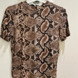Brun ormmönstrad topp från Zara - Säljer en kortärmad topp från Zara Trafaluc med coolt ormmönster i bruna och beige toner. Toppen har en rund halsringning och är figurnära i modellen. Perfekt för dig som vill sticka ut med ett djurmönster i din outfit.