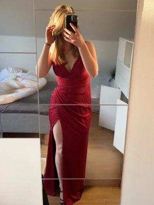 Röd balklänning med slits - Säljer en vinröd långklänning med djup v-ringning och smickrande omlottdetaljer i midjan. Klänningen har en hög slits framtill vilket ger en elegant och modern look. Perfekt för bal eller festliga tillfällen. Klänningen är endast använd en gång och är köpt på Proms and Weddings i Malmö med nypriset 2000 kr. Den har även vaddering vid bysten. Storleken är 6 vilket motsvarar en 34 men jag brukar ha M/38 och den passar mig perfekt!