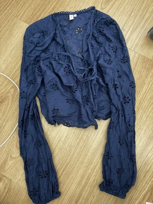 Nelly  blus med broderade detaljer mörkblå - Snygg mörkblå blus med långa ärmar och broderade hålmönster. Blusen har knytband framtill och vågig kant längs halsringningen. Tillverkad i ett lätt bomullstyg som ger en luftig känsla. Perfekt för dig som gillar romantiska detaljer och en avslappnad stil.