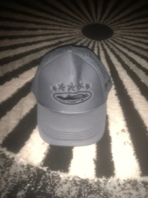 Grå meshkeps med broderad logga - Snygg grå keps med mesh på sidorna och bak, samt broderad logga framtill med fem stjärnor ovanför. Kepsen har böjd skärm och justerbar baksida för perfekt passform. Perfekt för dig som gillar streetwear och vill ha en stilren accessoar.