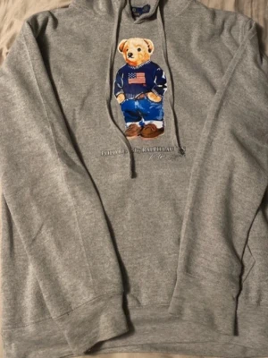 Grå hoodie Polo Ralph Lauren björn - Säljer en grå hoodie från Polo Ralph Lauren med den ikoniska björnen på bröstet, klädd i blå jeans och en tröja med amerikanska flaggan. Hoodien har huva med dragsko och ribbade muddar. Perfekt för dig som gillar klassisk streetwear med en twist. Bara att höra av sig vid frågor nyköpt för 2100!
