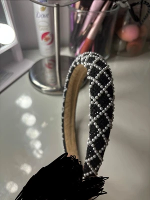 Svart diadem med pärlor - Snyggt svart diadem med ett rutmönster av vita pärlor. Diademet har en bred form och är klätt i ett mjukt tyg som ger en lyxig känsla. Perfekt accessoar för att lyfta vilken outfit som helst med en elegant touch.