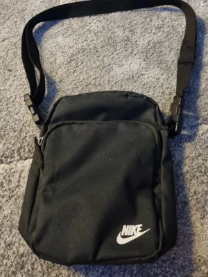 Svart axelväska med justerbart band - Snygg svart axelväska med enkel och stilren design. Väskan har ett justerbart axelband och praktisk form som passar perfekt för att bära det viktigaste. Tillverkad i slitstarkt material.