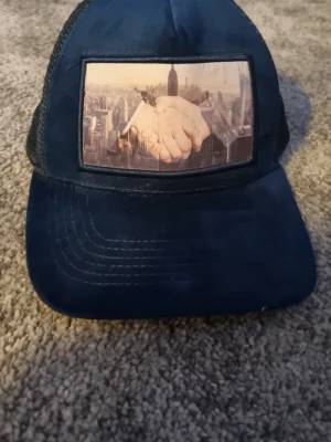 Marinblå truckerkeps från Gårda - Snygg marinblå truckerkeps från Gårda med mesh på sidorna och ett coolt tryck framtill som visar ett handslag mot en stadssilhuett. Justerbar baktill för perfekt passform. Perfekt för dig som vill sticka ut med en unik accessoar.