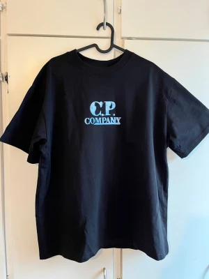 Svart C.P. Company t-shirt - Snygg svart t-shirt från C.P. Company med ljusblå logga tryckt på bröstet. Klassisk rund hals och korta ärmar. Perfekt för dig som gillar streetwear och vill ha en enkel men stilren look.
