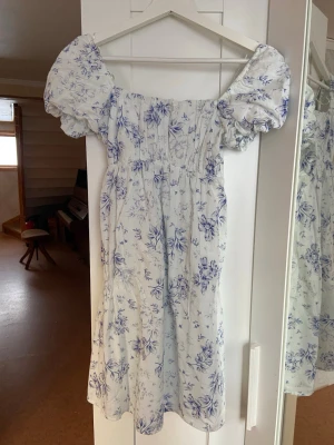 Vit blommig kortklänning med puffärm - Supersöt vit kortklänning med blått blommönster och puffiga kortärmar. Klänningen har en smockad överdel och fyrkantig urringning som ger en romantisk vibe. Perfekt för varma dagar och sommarhäng. (Bysten är inte stretchig) men jätteskön och flowig nertill! Har aldrig kommit till användning men jättefin. 🩵