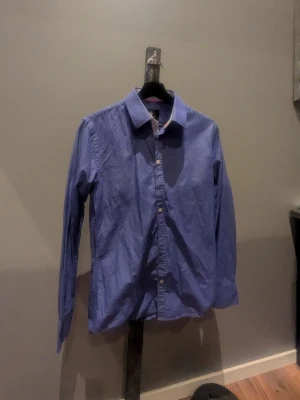 Blå skjorta med vit krage - Klassisk blå skjorta med vit kontrastkrage och vita knappar. Skjortan har lång ärm och normal passform, perfekt för en clean och stilren look. Tillverkad i bomull för skön känsla och enkel styling.