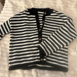 Randig svartvit kofta med knappar - Snygg kofta med svarta och vita horisontella ränder och guldiga knappar framtill. Koftan har rund halsringning, långa ärmar och ribbade muddar. Perfekt för en trendig och avslappnad look.