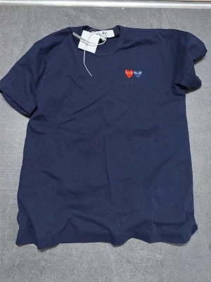 Mörkblå Comme des Garçons Play t-shirt - Snygg mörkblå t-shirt från Comme des Garçons Play med två små hjärtan i rött och blått broderade på bröstet. Klassisk passform och rund halsringning. Helt ny, tagg finns kvar och inga defekter. Köpt på Miinto, kvitto finns. Nypris 1000kr.