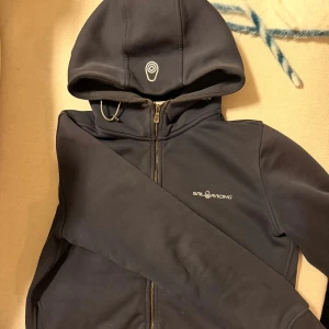 Marinblå hoodie från Sail Racing - marinblå hoodie från Sail Racing med dragkedja. Storlek ”L” för barn men passar en vuxen S. Nypris ligger på ca 1000-1500kr