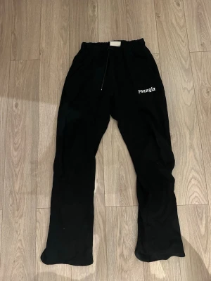 Svarta mjukisbyxor från YoungLA  - Svarta mjukisbyxor från YoungLA med tryck på ena benet och bakfickan. Byxorna har resår i midjan med snörning och raka ben. Perfekta för chill dagar och streetwear-vibe.