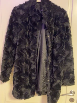 Svart pälsjacka med vågig struktur - Snygg svart pälsjacka med unik vågig struktur i materialet. Jackan har lång ärm, klassisk krage och knäppning framtill. Perfekt för dig som vill sticka ut med en trendig och mysig look. Fodrad insida för extra komfort.