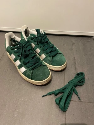 Gröna Adidas Campus sneakers  - Säljer ett par gröna Adidas Campus sneakers med klassiska vita ränder på sidorna och vit sula. Skorna har rund tå, snörning och kommer med extra gröna skosnören. Insidan är vit med Adidas-logga tryckt på sulan. + ett skosnöre😊