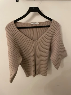 Beige ribbad topp från NA-KD - Säljer en beige ribbad topp från NA-KD med v-ringning och korta ärmar. Toppen har en loose passform och är tillverkad i ett mjukt stickat material som känns skönt mot huden. Perfekt att styla med jeans eller kjol för en trendig look.