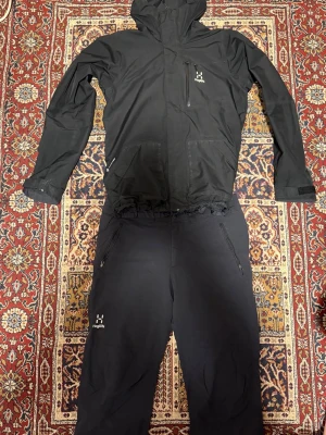 Svart vindjacka från Haglöfs - Säljer en svart vindjacka från Haglöfs med huva och dragkedja framtill och en haglöf byxor. Jackan har flera praktiska fickor, justerbara muddar och en sportig look. Tillverkad i slitstark polyester, perfekt för utomhusaktiviteter och vardagsbruk.