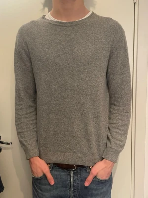 Grå stickad tröja i ullmix - En stilren grå stickad tröja med rund halsringning och långa ärmar. Tröjan har en klassisk passform och är tillverkad i mjuk ullmix som känns skön mot huden. Perfekt att matcha med jeans för en avslappnad look.