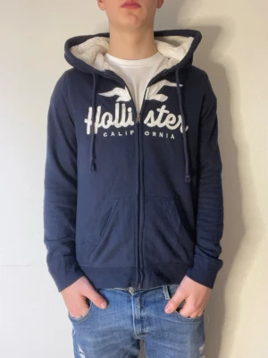 Mörkblå Hollister hoodie med dragkedja - Mörkblå hoodie från Hollister med vitt fluff inuti som är jätte skönt. Det är storlek medium men passar som small, skulle rekommendera för folk som är 170-175cm. Hör gärna av er vid minsta lilla fundering mvh// Partille closet