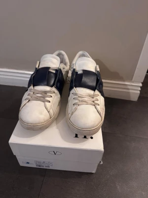 Valentino Open sneakers vit/marinblå 43 - Snygga Valentino Open sneakers i vitt skinn med en bred marinblå läderdetalj över vristen. Klassisk rund tå, platt sula och snörning framtill. Perfekta för dig som gillar exklusiva och stilrena sneakers med en unik touch.
