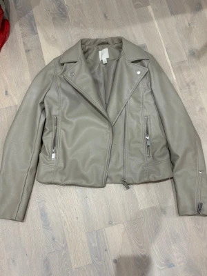 Beige skinnjacka med dragkedjor - Snygg beige skinnjacka med klassisk bikerkrage och silverfärgade dragkedjor framtill och vid ärmsluten. Jackan har två fickor med dragkedja och en croppad passform som ger en modern look.