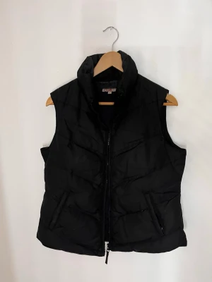 Svart väst - Svart dunväst från Cubus med hög krage och dragkedja framtill. Västen har två snedställda fickor med dragkedja och en stilren, enkel design som passar perfekt till lager-på-lager. Klassisk passform och quiltad struktur.