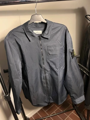 Stone Island - Snygg mörkblå overshirt från Stone Island med dragkedja framtill, bröstficka och klassisk patch på ärmen. Overshirten har en clean look och är tillverkad i ett lätt material som passar perfekt för lager-på-lager. Långärmad och med krage för en stilren vibe.