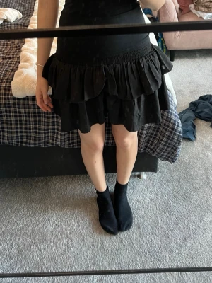 Svart volangkjol från Neo Noir - Supersnygg svart kortkjol från Neo Noir med bred resår i midjan och dubbla volanglager. Kjolen är gjord i 100% återvunnen polyester och har en flowy, luftig känsla som ger en cool look. Perfekt att styla med en enkel topp eller stickad tröja.