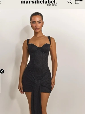 Svart kort klänning från Mars The Label - Supersnygg svart kort klänning med figurnära passform och breda axelband. Klänningen har en markerad byst, spetsliknande struktur och en knyt-detalj framtill som ger en extra cool touch. Perfekt för dig som vill sticka ut!
